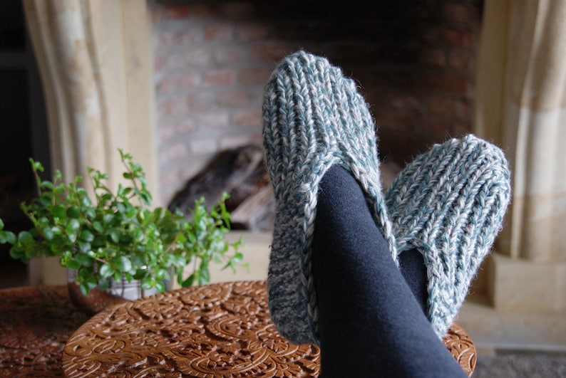 Puede incluir: Un par de zapatillas de punto acogedoras en un esquema de color azul y blanco. Las zapatillas son usadas por una persona que se relaja cerca de una chimenea.