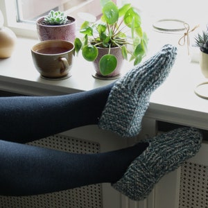 Puede incluir: Un par de calcetines de pantuflas de punto azul y blanco con las piernas de una persona que lleva medias negras descansando en un alf&eacute;izar de ventana. Hay plantas en macetas y una taza de t&eacute; en el alf&eacute;izar de la ventana.
