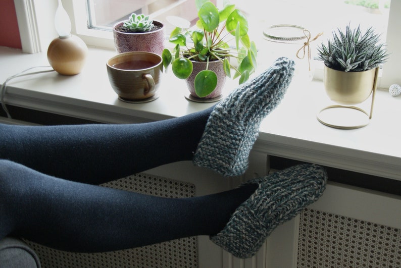 Puede incluir: Un par de calcetines de pantuflas de punto azul y blanco con las piernas de una persona que lleva medias negras descansando en un alf&eacute;izar de ventana. Hay plantas en macetas y una taza de t&eacute; en el alf&eacute;izar de la ventana.