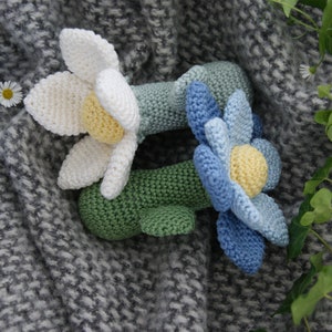 以下が含まれることがあります： 緑色の茎を持つ、2つの編みぐるみのおもちゃ。1つの花は黄色い中心を持つ白、もう1つの花は黄色い中心を持つ青です。