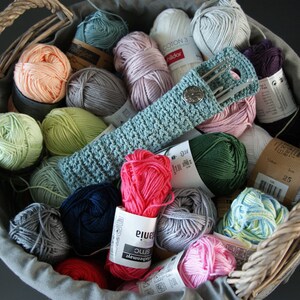 May include: A wicker basket filled with colorful yarn balls and a crocheted needle holder. The yarn is in various shades of pink, green, blue, and gray. The needle holder is light blue and has a button closure.