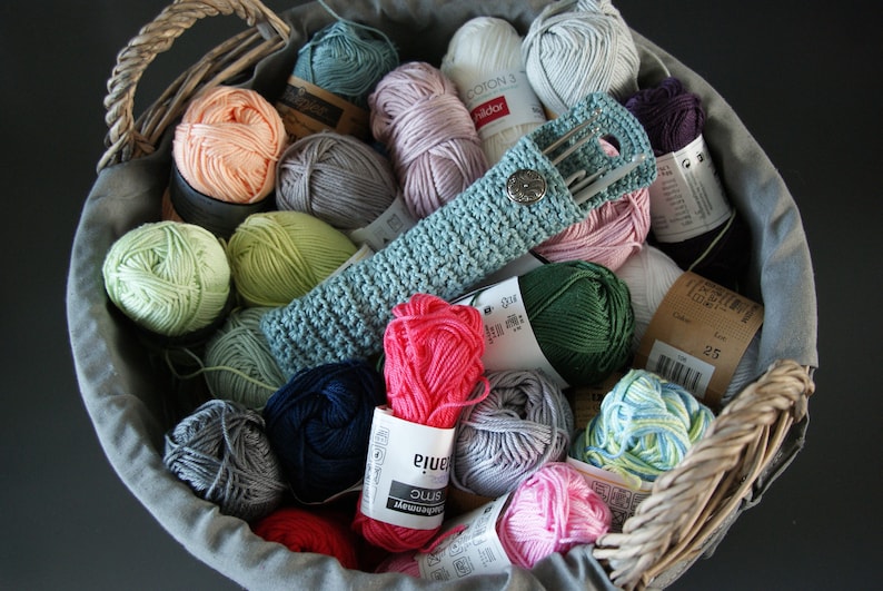 May include: A wicker basket filled with colorful yarn balls and a crocheted needle holder. The yarn is in various shades of pink, green, blue, and gray. The needle holder is light blue and has a button closure.
