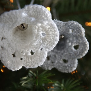 Puede incluir: Dos adornos de Navidad de ganchillo en forma de campanas, uno blanco y otro gris, colgados de un &aacute;rbol de Navidad.