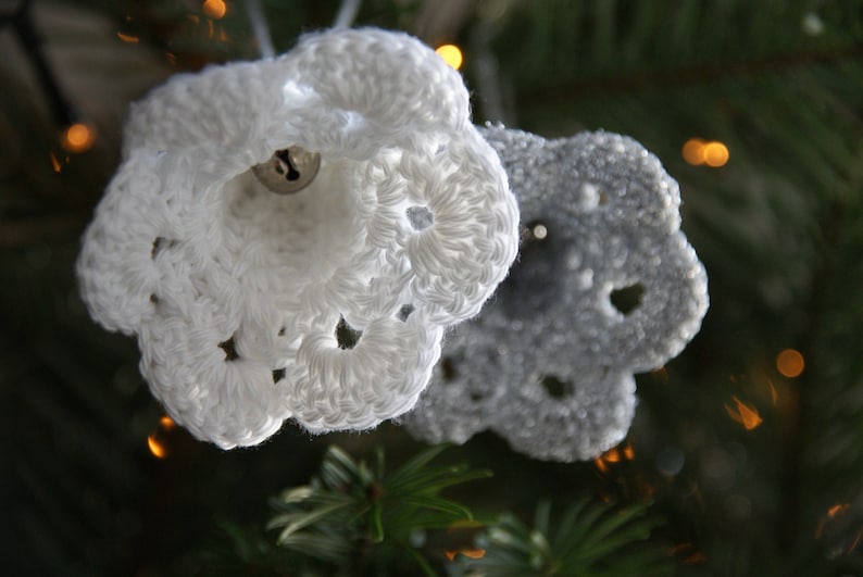 Puede incluir: Dos adornos de Navidad de ganchillo en forma de campanas, uno blanco y otro gris, colgados de un &aacute;rbol de Navidad.