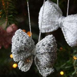 May include: Three crocheted bow ornaments hanging on a Christmas tree. One is white, one is silver, and one is pink.