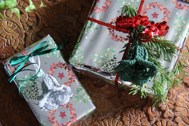 Puede incluir: Dos cajas de regalo plateadas envueltas con cinta verde y roja y decoradas con guirnaldas y estrellas verdes y rojas. Cada caja tiene un adorno de campana de ganchillo.