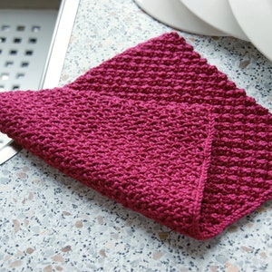 May include: A pink knitted dishcloth with a textured pattern. The dishcloth is folded and lying on a speckled worktop.