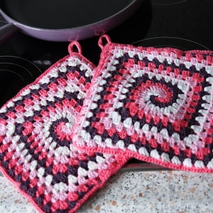 May include: Two crocheted pot holders in pink, white, and purple. The pot holders are square shaped and have a circular pattern in the center.