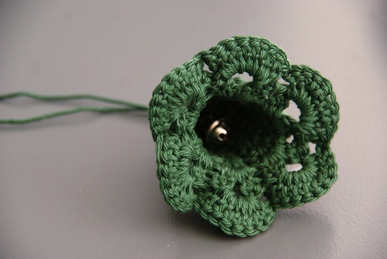 Puede incluir: Una campana de crochet verde con una campana de plata en su interior. La campana est&aacute; unida a una cuerda verde.