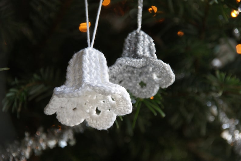 Puede incluir: Dos campanas de ganchillo, una blanca y otra plateada, colgando de un &aacute;rbol de Navidad.