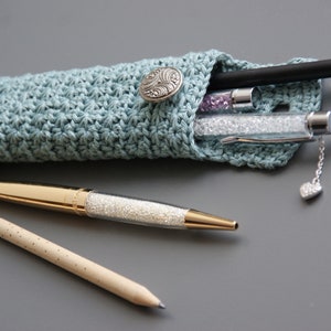 May include: A light blue crocheted pencil case with a silver button closure. The case is filled with pens and pencils, including a gold pen with a clear body and a black pen with a silver tip.