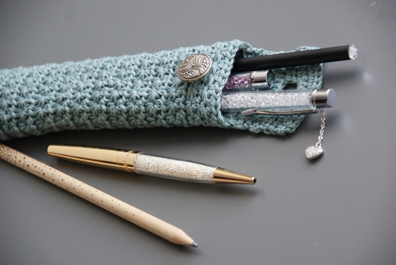 May include: A light blue crocheted pencil case with a silver button closure. The case is filled with pens and pencils, including a gold pen with a clear body and a black pen with a silver tip.