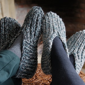 Puede incluir: Un par de zapatillas de punto acogedoras en un patr&oacute;n verde y blanco. Las zapatillas son usadas por una persona sentada cerca de una chimenea.