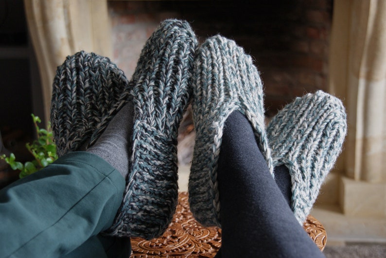 Puede incluir: Un par de zapatillas de punto acogedoras en un patr&oacute;n verde y blanco. Las zapatillas son usadas por una persona sentada cerca de una chimenea.
