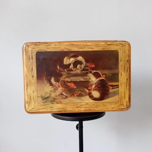Könnte beinhalten: Vintage rechteckige Blechdose mit einem Gemälde von drei Katzen um ein Goldfischglas. Die Dose hat ein Holzrahmendesign. Die Katzen sind braun, weiß und schwarz. Der Hintergrund ist in einem gedämpften, antiken Stil gehalten.