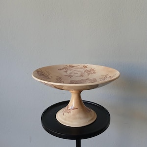 May include: A beige pedestal serving dish with a wide, shallow bowl and a short, rounded stem. The dish features a brown floral and architectural pattern. It is displayed on a small, black, round table.