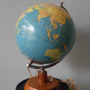 Op de afbeelding: Een vintage verlichte wereldbol met een blauwe oceaan en gele continenten. De wereldbol is gemonteerd op een houten voet met een lichtschakelaar en een netsnoer. De wereldbol wordt ondersteund door een metalen frame en staat op een zwarte standaard.