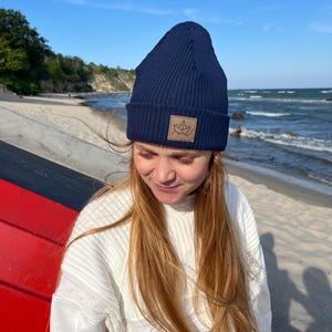 Könnte beinhalten: Marineblaue, gerippte Strickmütze mit einem kleinen braunen Lederaufnäher mit einem Blattmuster. Die Mütze wird von einer Person mit langen, hellbraunen Haaren getragen. Der Hintergrund zeigt einen Strand und das Meer unter blauem Himmel.