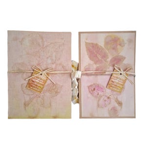 May include: Two handmade journals with botanical prints. The journals are tied with twine and have tags that read "ECOPRINT Artacarta". The covers are a light pink color with leaf imprints.