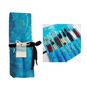 Op de afbeelding: Een blauwe batik-patroon kunstenaarspenseelrol met een zwarte striksluiting. De rol is vastgebonden met een zwart lint en bevat diverse penselen. De rol is gemaakt van stof en heeft een label met de tekst "Aricarla".