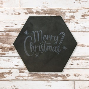 May include: A black, hexagonal sign featuring the text "Merry Christmas" in a cursive script, decorated with snowflakes and a candy cane. The sign is placed on a white, distressed wooden surface.