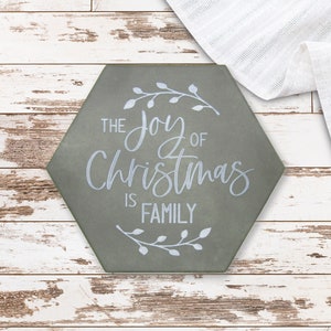 May include: A hexagonal sign in muted green, featuring the white script "THE JOY OF CHRISTMAS IS FAMILY" and floral accents. It rests on a distressed white wooden surface, with white fabric draped nearby. The sign is a festive decoration.