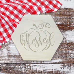 May include: A light-colored, hexagonal wooden sign with the word "blessed" in a cursive font, and a pumpkin design. The sign is on a rustic, white-washed wooden surface, with a red and white checkered cloth in the background.