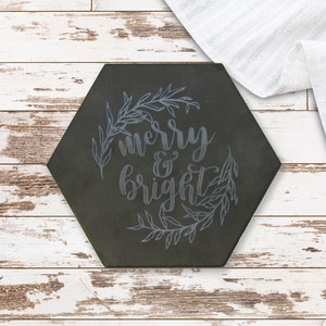 May include: A dark grey, hexagonal tile featuring the words "merry & bright" in cursive, framed by a leafy wreath. It sits on a distressed white wooden surface, with white fabric in the background.