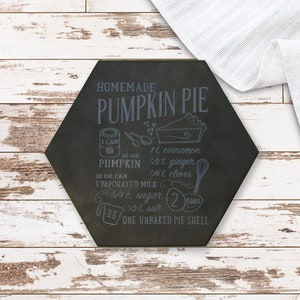 Peut inclure: Une tuile décorative hexagonale noire avec les mots "HOMEMADE PUMPKIN PIE" et une recette de tarte à la citrouille. La tuile est sur une surface en bois blanc vieilli avec un tissu blanc.
