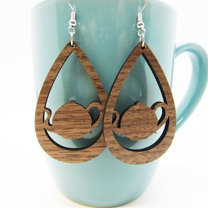 May include: A pair of wooden teardrop-shaped earrings with a cutout silhouette of a teapot. The earrings are brown and have a natural wood grain finish.