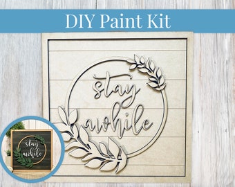 May include: DIY paint kit featuring a wooden sign with the words "stay awhile" in a circle with leafy branches. The sign is unfinished and ready to be painted.