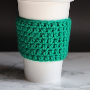 May include: A white to-go coffee cup with a green crocheted cozy.