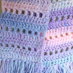 Puede incluir: Una bufanda de ganchillo con un degradado de colores rosa, morado y azul. La bufanda tiene flecos en la parte inferior y está hecha con un patrón de punto sencillo.