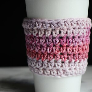 May include: A crocheted coffee cup cozy in shades of pink and purple. The cozy is made with a simple stitch pattern and has a gradient of color.