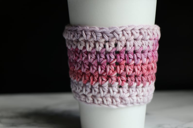 May include: A crocheted coffee cup cozy in shades of pink and purple. The cozy is made with a simple stitch pattern and has a gradient of color.