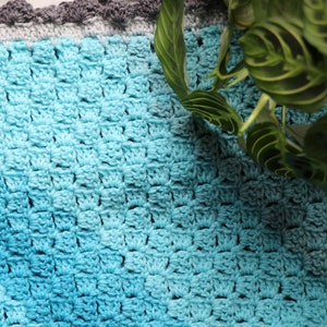 Peut inclure: Une couverture en crochet bleu et gris avec un dégradé de couleur. La couverture a un bord festonné et est réalisée avec un point texturé.
