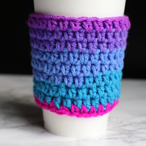 May include: A white coffee cup with a crocheted cozy in shades of purple, blue, and pink. The cozy is made of multiple stripes of different colors.