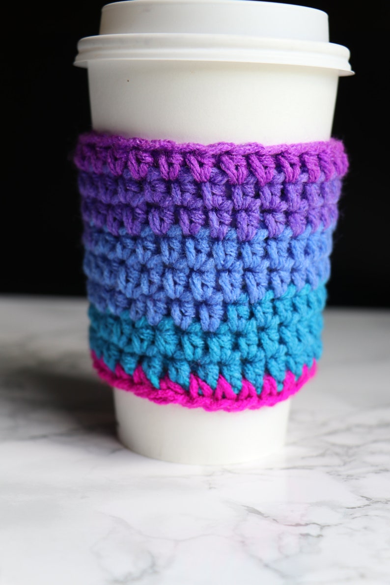 May include: A white coffee cup with a crocheted cozy in shades of purple, blue, and pink. The cozy is made of multiple stripes of different colors.