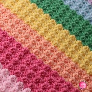 Può includere: Una coperta all'uncinetto arcobaleno con un motivo a punto testurizzato. La coperta è realizzata in tonalità di rosa, arancio, giallo, verde e blu.