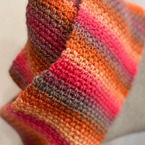 Puede incluir: Primer plano de una bufanda de ganchillo en tonos naranja, rosa y marrón. La bufanda está doblada por la mitad y se aprecia la textura de los puntos de ganchillo.