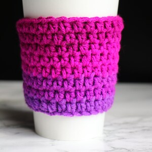 May include: A crocheted coffee cup cozy in shades of pink and purple. The cozy is made with a simple stitch pattern and is perfect for keeping your hands warm while enjoying your favorite beverage.