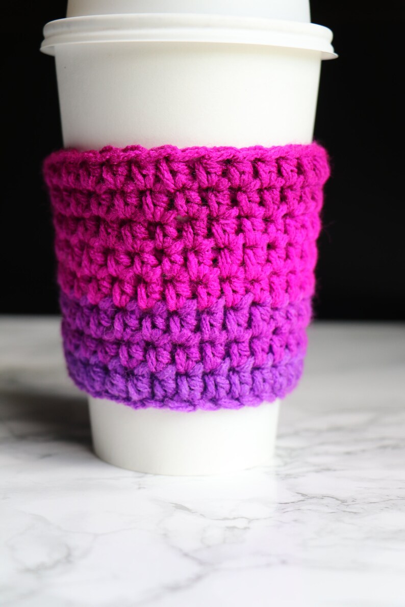 May include: A crocheted coffee cup cozy in shades of pink and purple. The cozy is made with a simple stitch pattern and is perfect for keeping your hands warm while enjoying your favorite beverage.