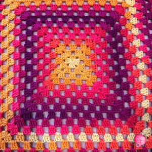Könnte beinhalten: Eine quadratische, handgefertigte Häkeldecke mit einem lebendigen Farbverlauf. Die Mitte ist ein warmes Gelb, das über Orange, Rosa und Lila zu einem hellbeigen Rand übergeht. Das Granny-Square-Muster ist deutlich sichtbar.