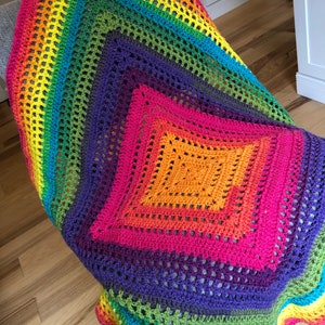 May include: A rainbow crochet blanket with a square pattern in the center. The blanket is made with different colors of yarn, including red, orange, yellow, green, blue, indigo, and violet.