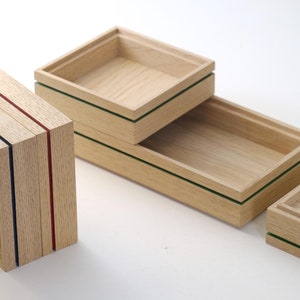 Op de afbeelding: Vier houten dozen met verschillende gekleurde randen. De dozen zijn op elkaar gestapeld. De bovenste doos heeft een groene rand, de tweede doos heeft een groene rand, de derde doos heeft een rode en blauwe rand, en de onderste doos heeft een groene rand.