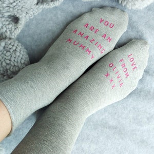 May include: Two pairs of gray socks with pink glitter. One sock says 'You are an amazing mummy' and the other sock says 'Love from Olivia xxx'.