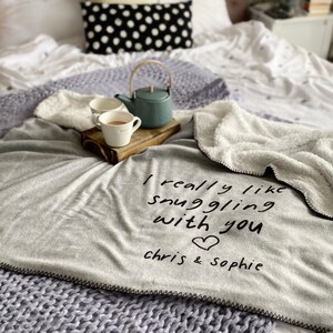 May include: A gray fleece blanket with the text "I really like snuggling with you" and a heart symbol. The text "chris & sophie" is below the heart. Two mugs and a teapot are on a wooden tray in the background.