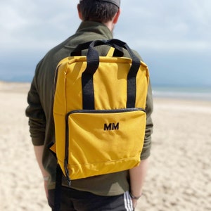 Puede incluir: Una mochila de lona amarilla con correas negras y una cremallera negra. La mochila tiene un monograma bordado en negro "MM" en la parte delantera.