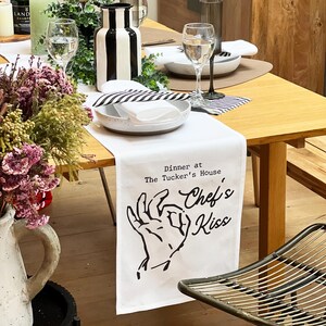 May include: White table runner with black stripes and the text "Dinner at The Tucker's House Chef's Kiss" with a black and white illustration of a hand giving a thumbs up.