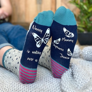 May include: A pair of blue and teal socks with a pink stripe at the top. The socks have white butterflies and text that reads "Nanny is watching over us", "Mummy", "Stephanie", and "Jessica".